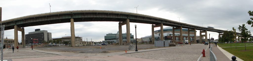  /><span>The aging and fracture critical Buffalo Skyway bridge looms over the Buffalo Harbor project site. (Image: Wayne Senville, Planning Commissioners Journal, www.plannersweb.com)</span></p>
<p><strong>Like a number of other 1950’s-era, aging elevated highways that are (or have already) reached the end of their designed lifespans, the elevated Buffalo Skyway bridge in New York could be the latest to be replaced with a more people-centric design that better connects the city to its waterfront. Smart Growth America’s CEO Calvin Gladney has been selected to join a panel of other notable experts to judge a state-chartered design competition about its future.</strong></p>
<p>Built during the 1950’s at a height that allowed lake ships to pass underneath back when Buffalo was a busy port city, today’s Buffalo Skyway is a relic from a different age and a perpetual headache for residents and city/state officials.</p>
<p>It blocks downtown from its waterfront, it occupies valuable yet underutilized waterfront land, and requires seemingly never-ending expensive construction just to keep the structurally deficient and fracture critical bridge limping along. A recent state proposal in 2014 to modestly rehabilitate and maintain the bridge as-is would cost $42 million over a 20-year period, <a href=