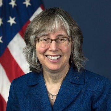 Barbara McCann headshot wearing glasses, a blue blazer, in front of an American Flag over her left shoulder