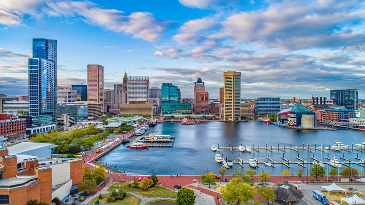 A vivid look at Baltimore's inner harbor. A wide red sidewalk curves around the waterfront and tall buildings rise in the background.